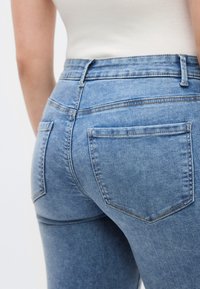 Vékony szabású világoskék farmernadrág magas derékrésszel, két hátsó zsebbel és enyhe kifakulással. Az anyag enyhén kopott hatású, varrási részletekkel.