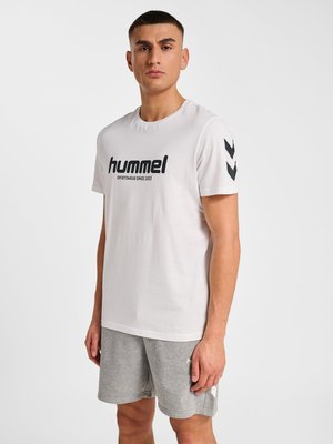 Mann trägt weißes Hummel-T-Shirt mit schwarzem Logo und grauen Shorts, steht vor einfachem hellem Hintergrund.