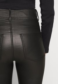 Gros plan d'une personne portant un pantalon noir taille haute en simili cuir avec des poches arrière visibles et un haut noir à manches longues.