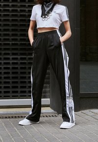 Svarta träningsoveraller med vita sidostripes, tryckknappar och Adidas-logotyp. Matchas med en kort vit tröja och vita sneakers.