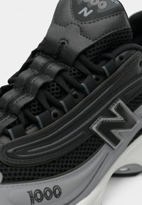Close-up of black and gray New Balance 1000 sneaker showing mesh fabric, leather details, laces, and logo on midsole and tongue.