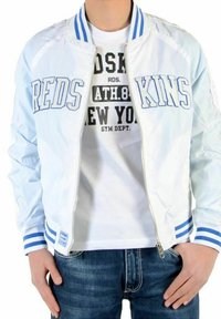 Lyseblå bomberjakke med hvidt og blåt stribet kantbånd, prydet med teksten "REDS KINS" på forsiden, båret over en hvid t-shirt.