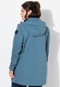 Chaqueta azul impermeable con capucha, mangas largas y botones laterales. Tejido texturizado con un acento negro en el hombro.