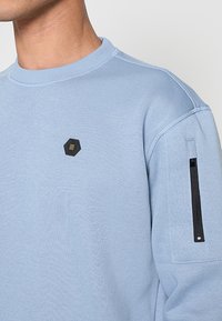 Lichtblauwe sweatshirt met een ronde hals, textuurstof, een zeshoekig zwart logo op de borst en een zwarte ritssluitingzak op de mouw.