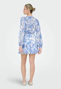 Femme aux cheveux blonds coiffés en chignon, portant une robe à manches longues bleue et blanche à motif floral et des chaussures beige sans lacets, debout, tournée de dos.