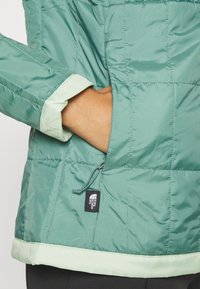 Zelená izolační bunda s hladkou texturou, obtaženými manžetami a kontrastním světle zeleným lemem. Obsahuje štítek s logem na bočním švu.