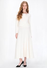 Ensemble deux pièces texturé couleur crème, composé d'un cardigan court avec un détail en nœud et d'une longue jupe fluide, associé à des talons aiguilles noirs.