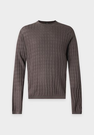 Homme portant un pull marron texturé, un pantalon large noir et des chaussures noires, debout avec une main dans la poche sur un fond uni.