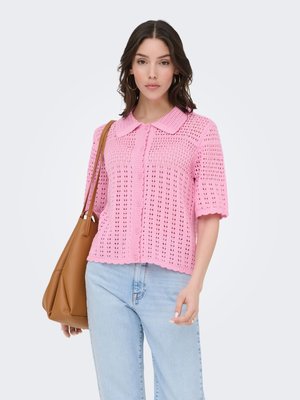 Femme portant un haut en maille rose à manches courtes avec col, un jean bleu clair, des boucles d’oreilles créoles dorées et un grand sac à bandoulière en cuir marron.