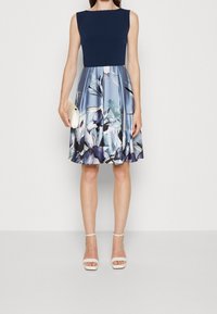 Ärmelloses Kleid mit einem marineblauen Oberteil und einem hellblauen Rock, der mit floralen Mustern in Lila und Grün versehen ist, kombiniert mit weißen Sandalen.
