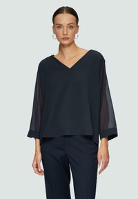 Dunkelblaue Bluse mit V-Ausschnitt, dreiviertellangen, transparenten Ärmeln und einem lockeren Schnitt, kombiniert mit strukturierten dunklen Hosen.