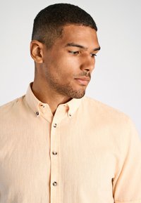 Knopenblouse in licht perzikkleurige stof met een textuurafwerking, korte mouwen en metalen knopen langs de sluiting en de kraag.