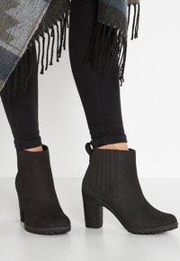 Bottes noires en daim à la cheville avec un talon texturé, des panneaux latéraux élastiques et une languette. Portées avec un jean skinny noir et un châle à franges.