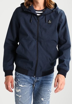 Person iført en mørkeblå hættetrøje med lynlås og sorte manchetter, kombineret med slidte lyseblå jeans.