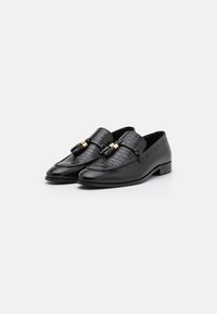 Mocasines negros con un patrón de cocodrilo texturizado, con una correa frontal acentuada con herrajes dorados. Exterior de cuero, diseño de tacón plano.
