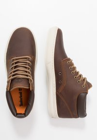 Bruine leren hoge sneakers met tan veters, een crèmekleurige rubberen zool en een versterkte neus, met een logo op de tong en gestikte accenten.
