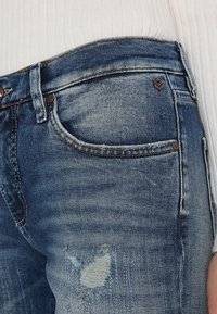 Siniset farkut, joissa on rento istuvuus, etupussissa on kulunut kohta ja ruskeat metalliosat. Kangas on tekstuuripintainen.