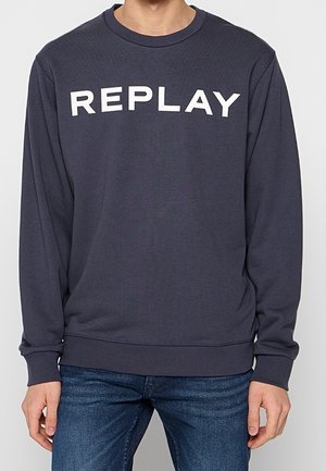 Donkergrijze sweater met lange mouwen en "REPLAY" in grote witte hoofdletters op de borst, gedragen met een blauwe jeans.