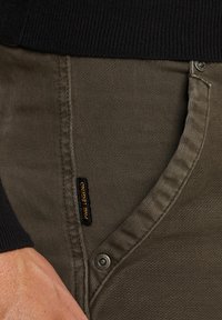 Pantalons kaki en tissu texturé, avec une étiquette de logo discrète et une fermeture à bouton en métal. La taille est sombre et côtelée.