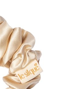 Satynowa gumka do włosów w jasnym złocie o gładkiej fakturze. Zawiera metkę z napisem "lesilque" w kolorze pomarańczowym.