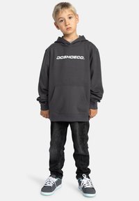Grijze hoodie met een wit "DCSHOECO." logo, voorzak, geribbelde manchetten; gecombineerd met donkere jeans en grijze sneakers met teal accenten.
