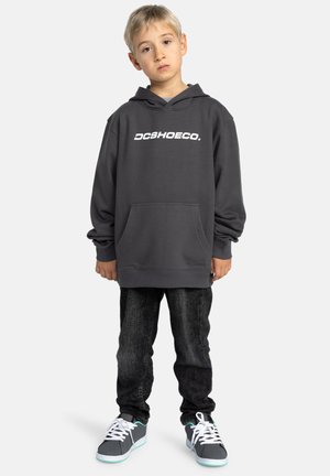 Grijze hoodie met een wit "DCSHOECO." logo, voorzak, geribbelde manchetten; gecombineerd met donkere jeans en grijze sneakers met teal accenten.