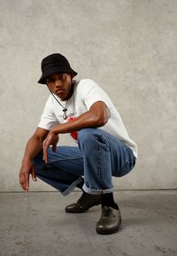 Sapatos de couro castanho com um acabamento suave, usados com jeans de ganga dobrados e uma t-shirt branca com uma estampa. Um chapéu bucket preto completa o visual.