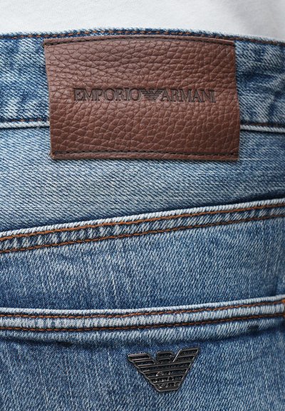 Ruda odinė etiketė su įspaustu "EMPORIO ARMANI" ant mėlyno džinsinio audinio, išsiskirianti kontrastinga oranžine siūle ir juodu logotipo akcentu.