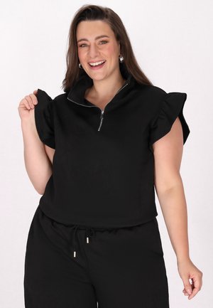 Top corto de manga corta negro con cuello con cremallera y tela texturizada. La modelo sostiene la manga con volantes mientras lleva unos pantalones negros a juego.