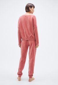 Ensemble de vêtements d'intérieur en velours rose composé d'un pull ample à texture côtelée et d'un pantalon assorti coupé près des chevilles, pieds nus visibles.