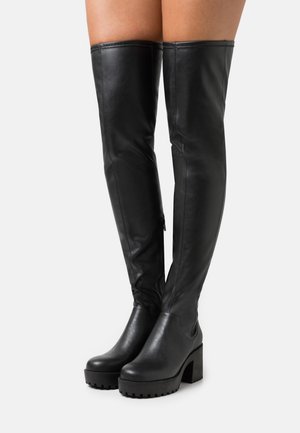 Bottes en cuir noires montantes jusqu'aux cuisses avec talons hauts épais portées sur des jambes croisées sur un fond clair uni.