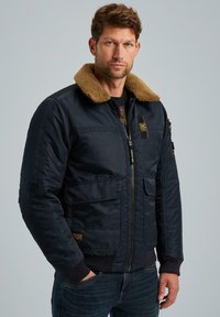 Marineblaue Bomberjacke mit braunem Kunstfellkragen, Reißverschluss, mehreren Fronttaschen sowie gerippten Bündchen und Saum.