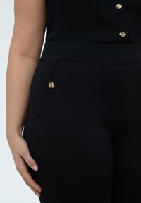 Primer plano de una persona con pantalones y parte superior negras, con botones decorativos dorados cerca del bolsillo y en el pecho, con la mano descansando al costado.