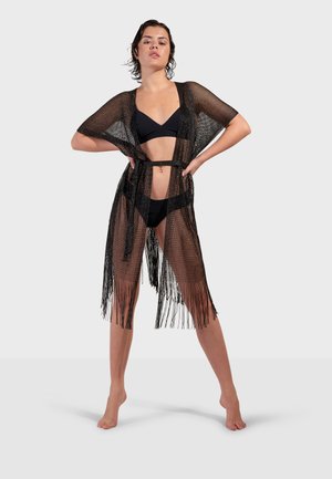 Femme debout pieds nus portant un bikini noir et un paréo noir transparent à franges, mains sur les hanches, devant un fond clair uni.