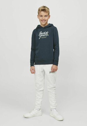 Felpa blu scuro con logo bianco, abbinata a pantaloni della tuta grigio chiaro e scarpe da ginnastica bianche. Tessuto morbido, vestibilità comoda e cappuccio con coulisse.