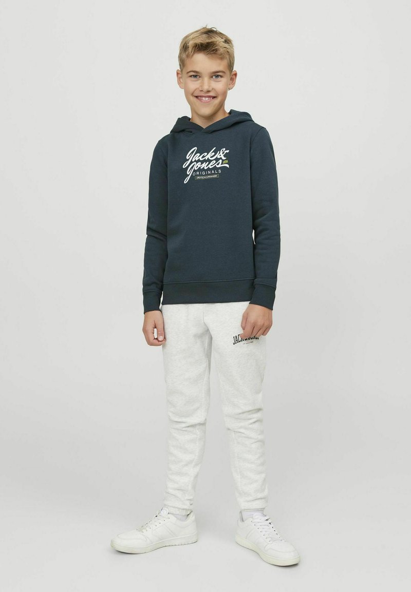 Felpa blu scuro con logo bianco, abbinata a pantaloni della tuta grigio chiaro e scarpe da ginnastica bianche. Tessuto morbido, vestibilità comoda e cappuccio con coulisse.