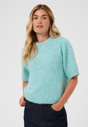 Femme aux longs cheveux blonds portant un pull doux bleu clair à manches courtes et un jean bleu foncé, debout avec une main dans la poche.