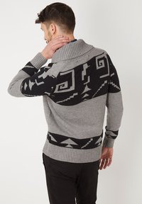 Suéter de punto gris con un gran cuello, que presenta patrones geométricos negros a lo largo de los hombros y la parte inferior. Puños y dobladillo acanalados.