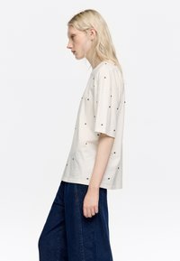 Koszulka z krótkim rękawem, w kolorze kremowym, wykonana z bawełny, z małymi czarnymi zdobieniami. Luźny krój i okrągły dekolt. Połączona z ciemnoniebieskimi spodniami o szerokich nogawkach.