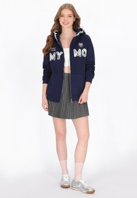 Sudadera azul marino con cremallera y letras blancas con patrón, falda plisada gris, zapatillas plateadas y calcetas de red. La capucha tiene un ribete fruncido.