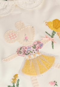 Stickerei einer Maus in einem blumigen Kleid und einem gelben Tüllrock. Der Hintergrund ist cremefarben, mit dekorativen Blumen, die in der Nähe gestickt sind.
