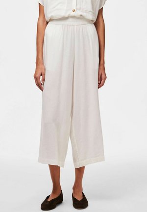 Fehér, bő szárú nadrág, könnyű anyagból készült. Rugalmas derékpánttal és laza fazonnal, feltekert szegéllyel. Barna slip-on cipőkkel viselve.