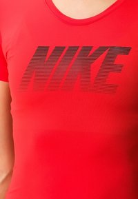 Punainen Nike-aktiivivaatteiden paita, jossa on pyöreä pääntie, mustalla logolla, jossa on vaakasuoria raitoja. Valmistettu sileästä, joustavasta kankaasta.