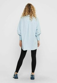 Camicia oversize azzurra con maniche a palloncino, polsini abbottonati, dettaglio di piega sul retro, abbinata a leggins neri e scarpe da ginnastica blu.