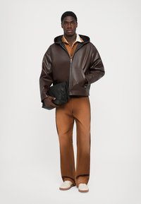 Brun læderjakke med hætte og lynlås, parret med en brun skjorte og brede brune bukser, mens der holdes en sort clutch.