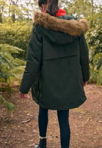 Parka verte avec une capuche en fausse fourrure, détail de fermeture éclair et poches latérales. Superposée sur un intérieur rouge, portée avec des leggings noirs.