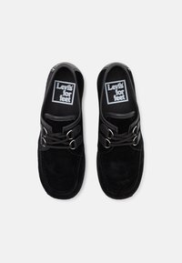 Chaussures slip-on en daim noir avec deux boucles métalliques et des lacets noirs. Intérieur marqué du logo "Levi's for feet" en texte blanc.