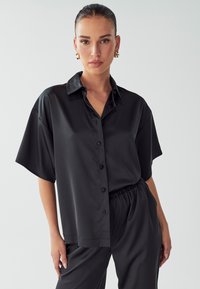 Camicia nera di satin a maniche corte con un design a bottoni, dotata di colletto classico e texture liscia. Abbinata a pantaloni coordinati.