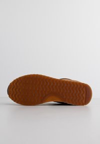 Semelle de chaussure avec une bande de roulement en caoutchouc marron comportant des rainures ondulées et un talon plat sur un fond clair uni.