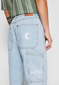 Pantalon en denim bleu clair avec une poche arrière ornée d'un logo "K" brodé en blanc et d'une étiquette en cuir de marque au niveau de la taille. Texture douce.